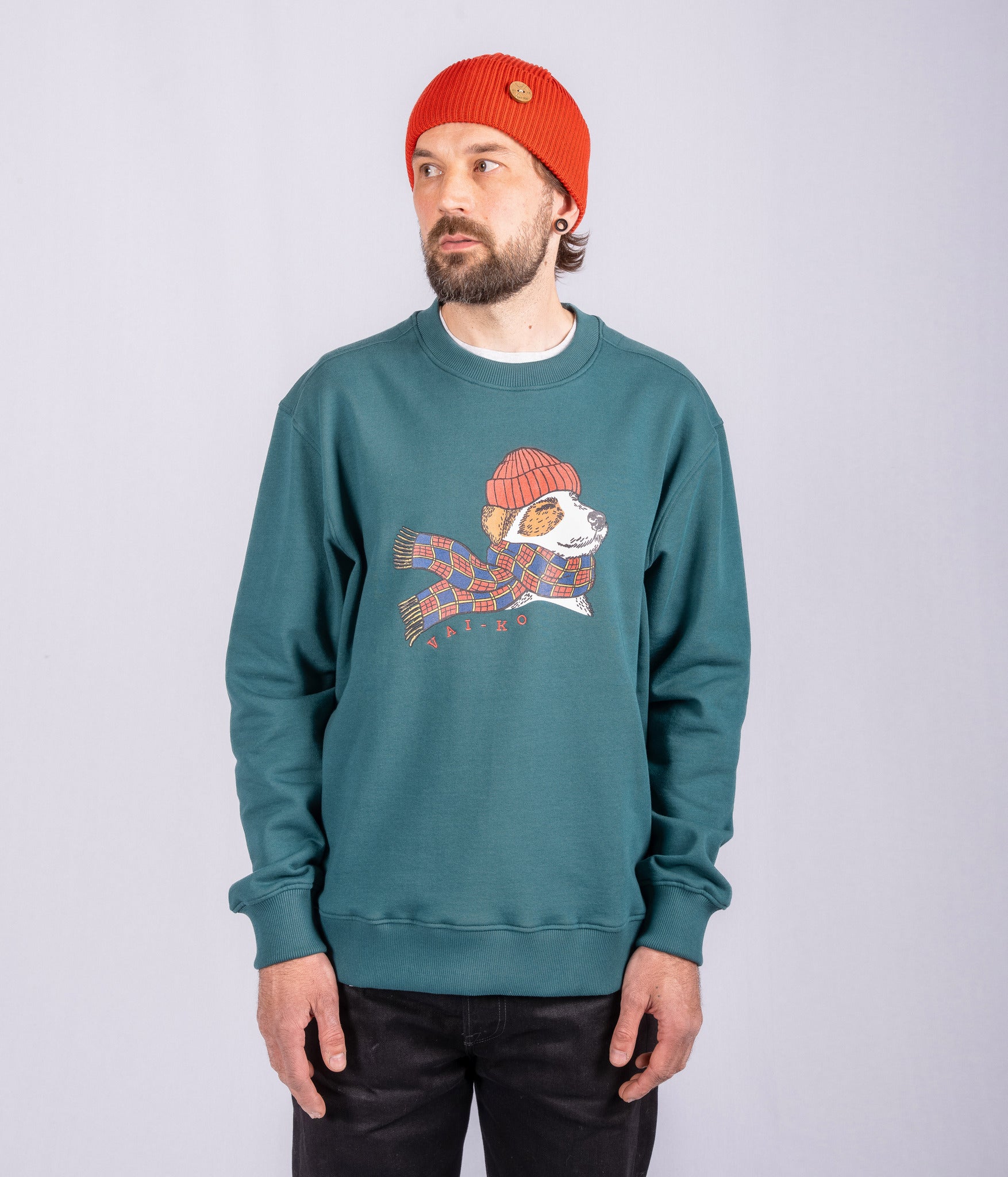 Man in teal sweatshirt with dog graphic, wearing red beanie, standing against a plain light background.