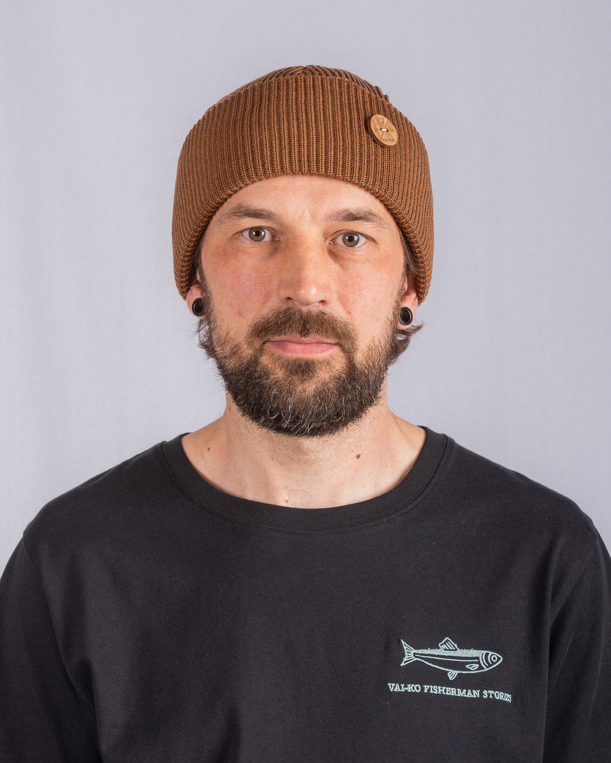 Man with a brown beanie and black shirt stands against a plain light gray background, looking at camera.