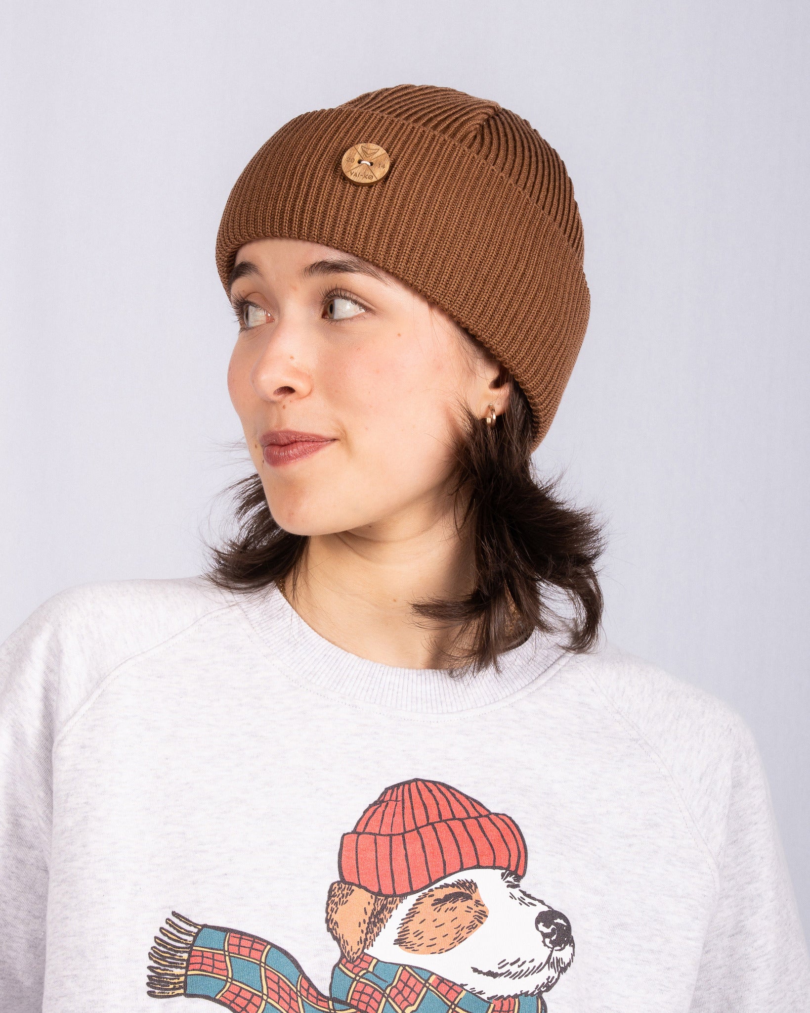 Woman in a brown knit beanie looks sideways, wearing a sweater with a dog in a red hat and scarf print.