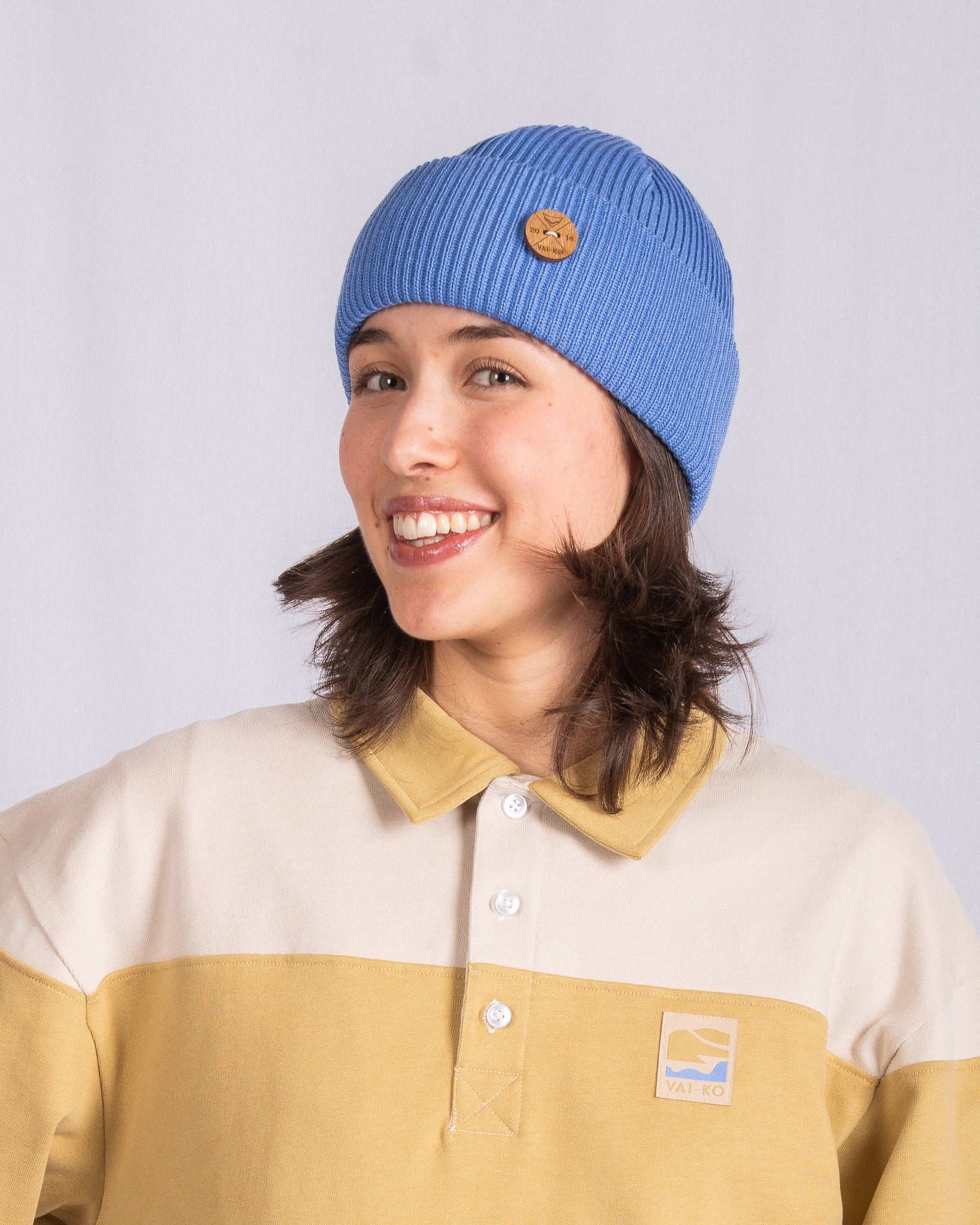 Smiling person wearing a blue beanie and a beige-and-yellow collared shirt against a plain background.