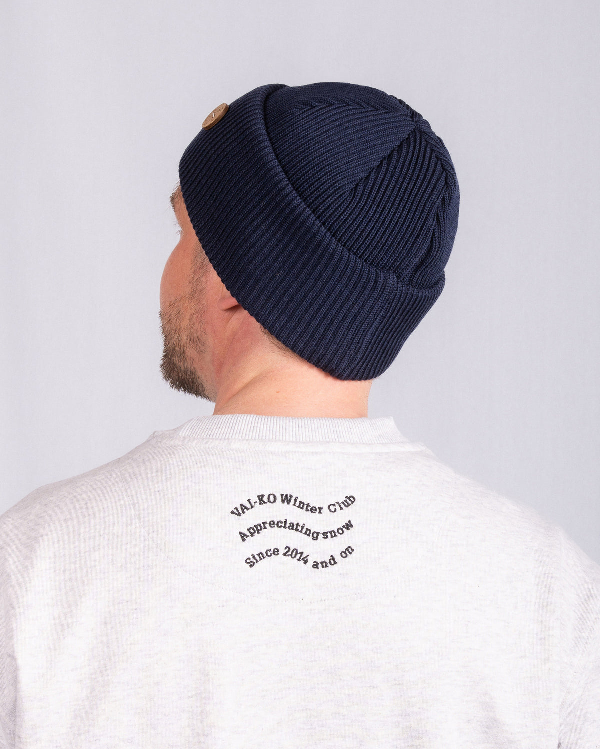 Man in a navy knit beanie and a light sweatshirt with text printed on the back, facing away.