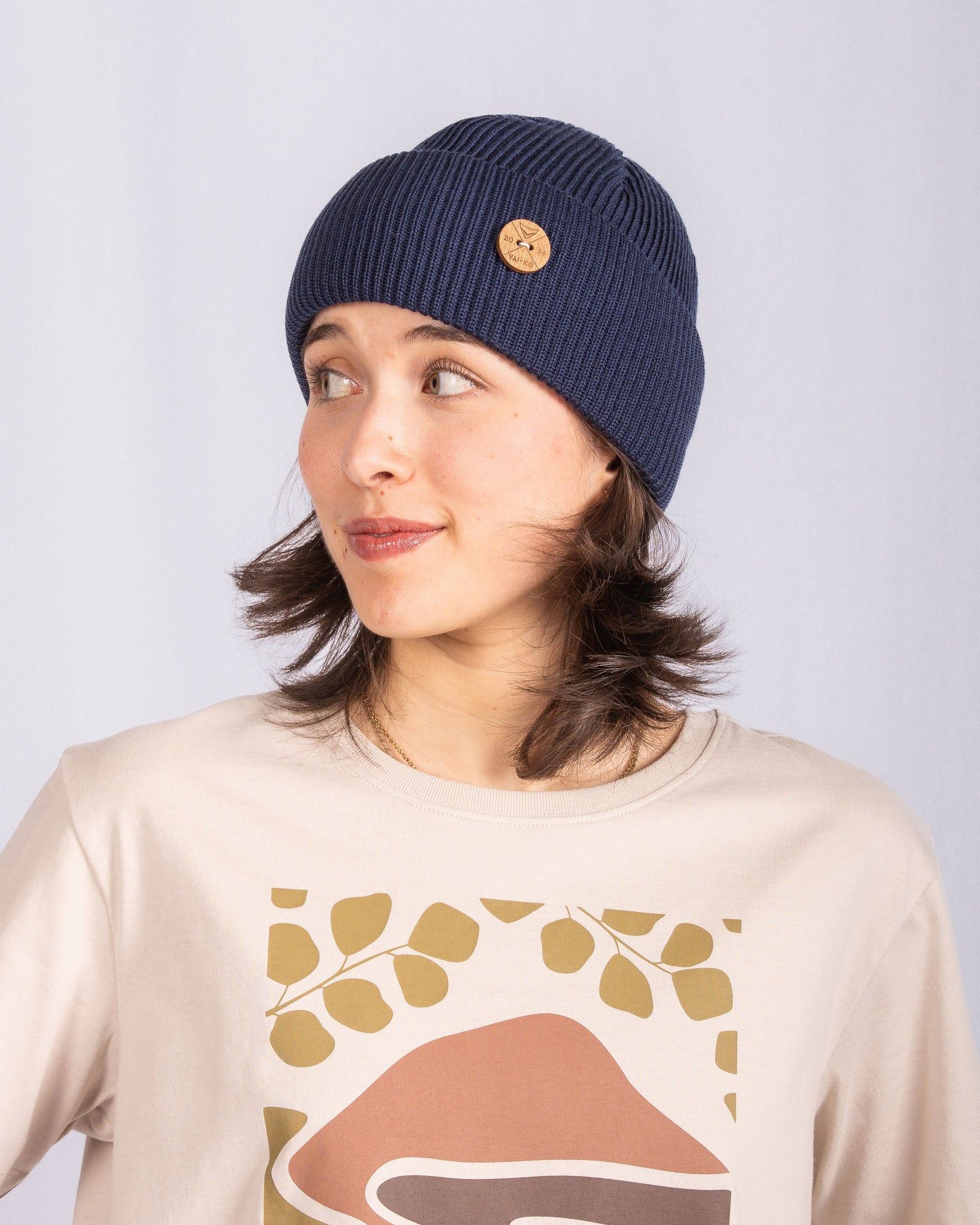 Woman in a blue knit beanie and printed beige shirt, looking to her left, against a plain light background.