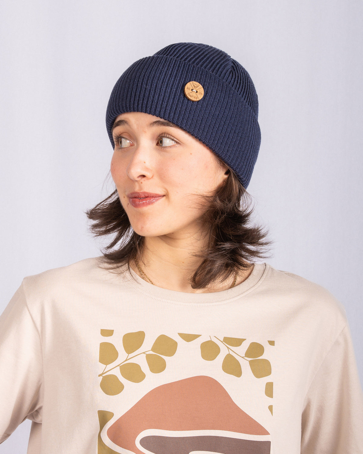 Woman in a blue knit beanie and printed beige shirt, looking to her left, against a plain light background.
