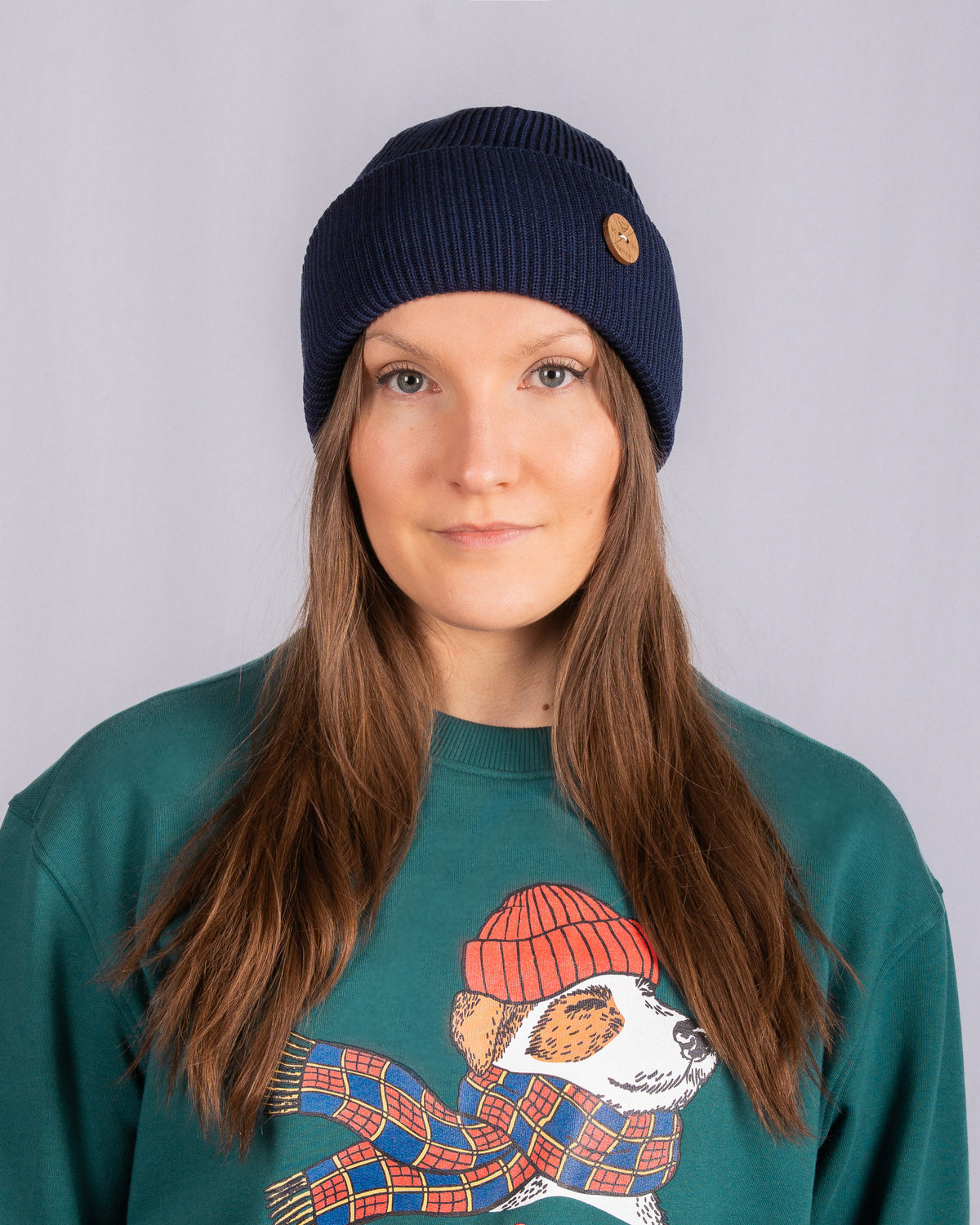Woman with long brown hair wearing a navy beanie and green sweater featuring a dog in a red hat and scarf.