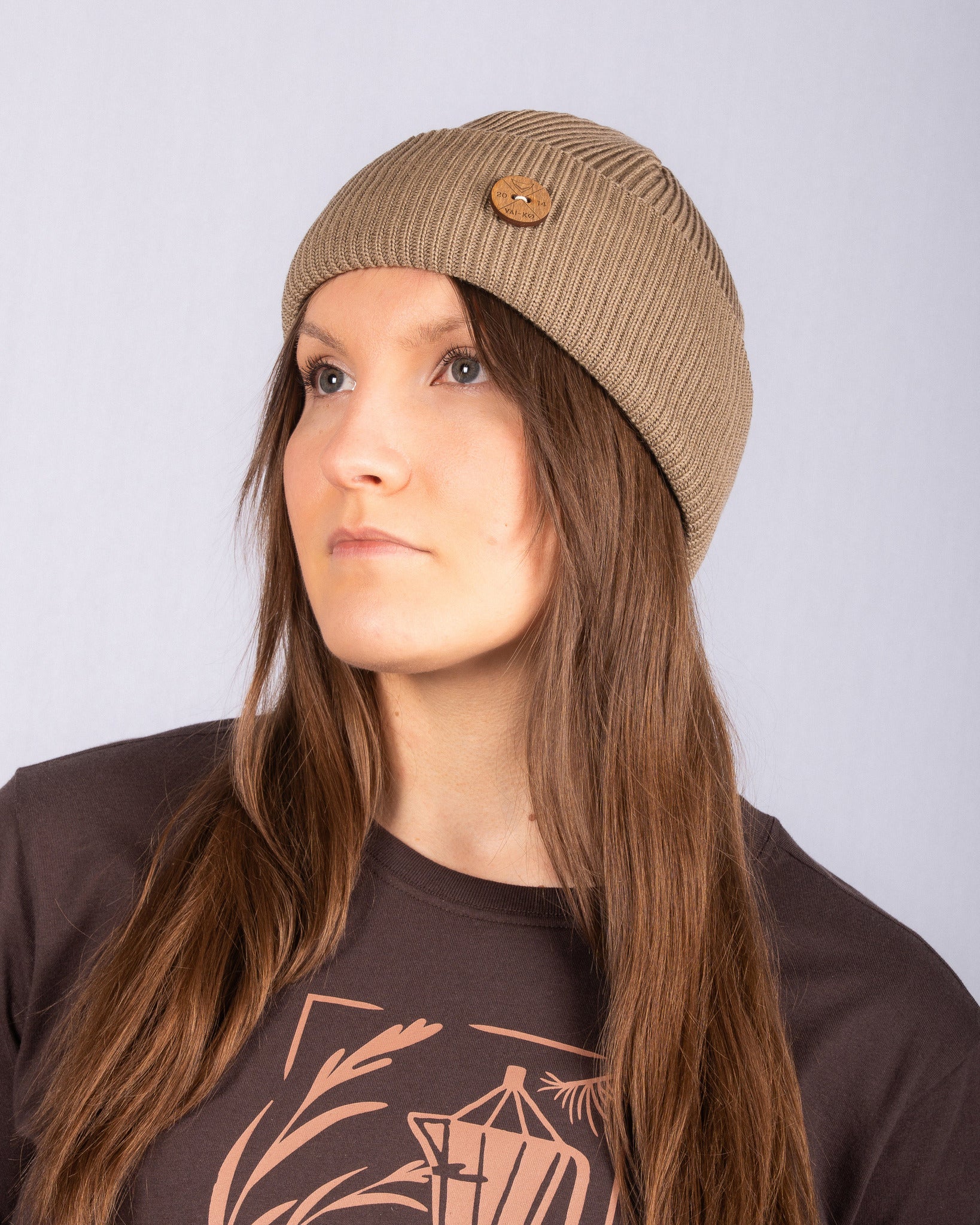 Woman with long brown hair wearing a tan knit beanie and a dark shirt, looking to the left.