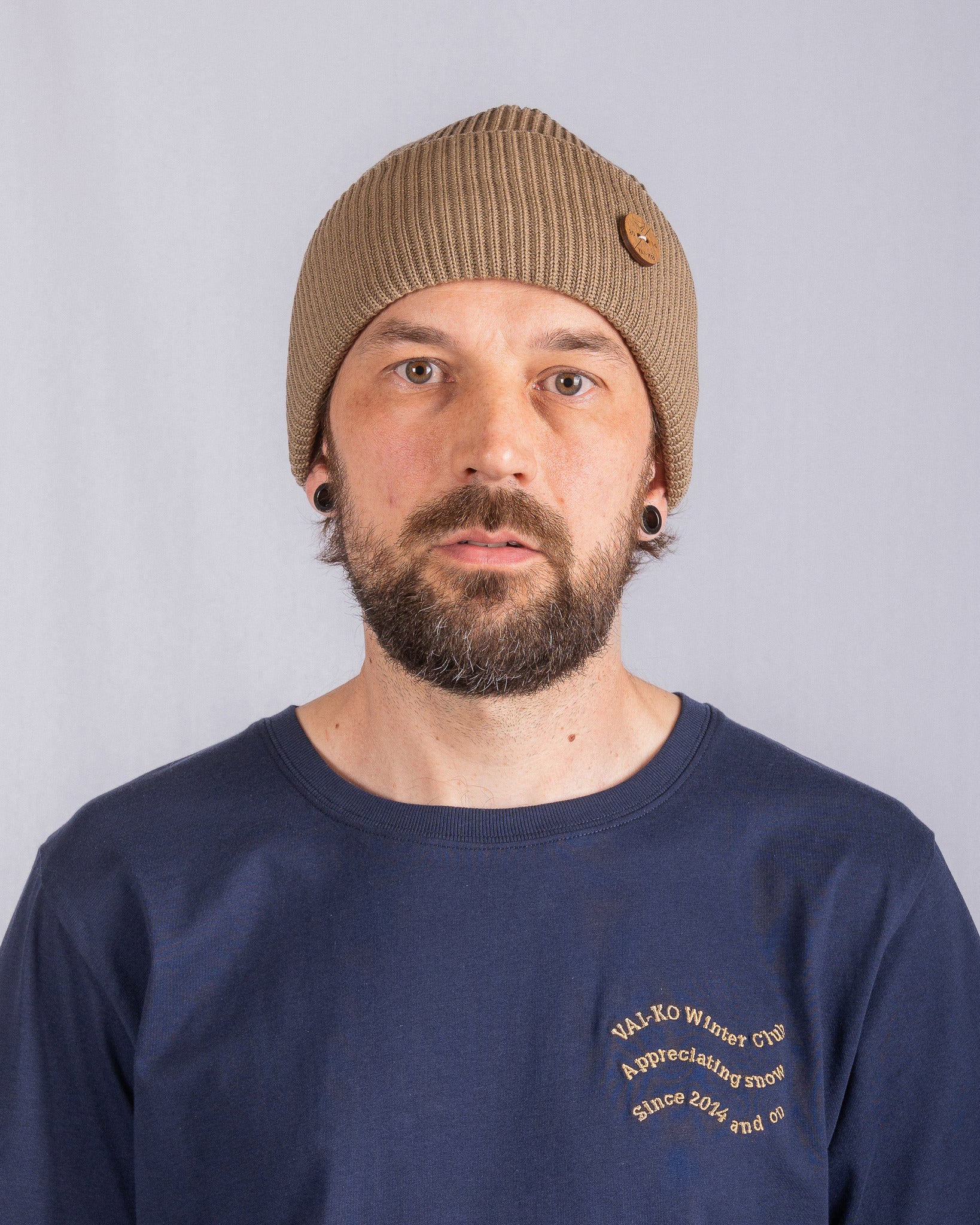Man with a brown beanie and blue shirt stands against a plain light background, looking at the camera.