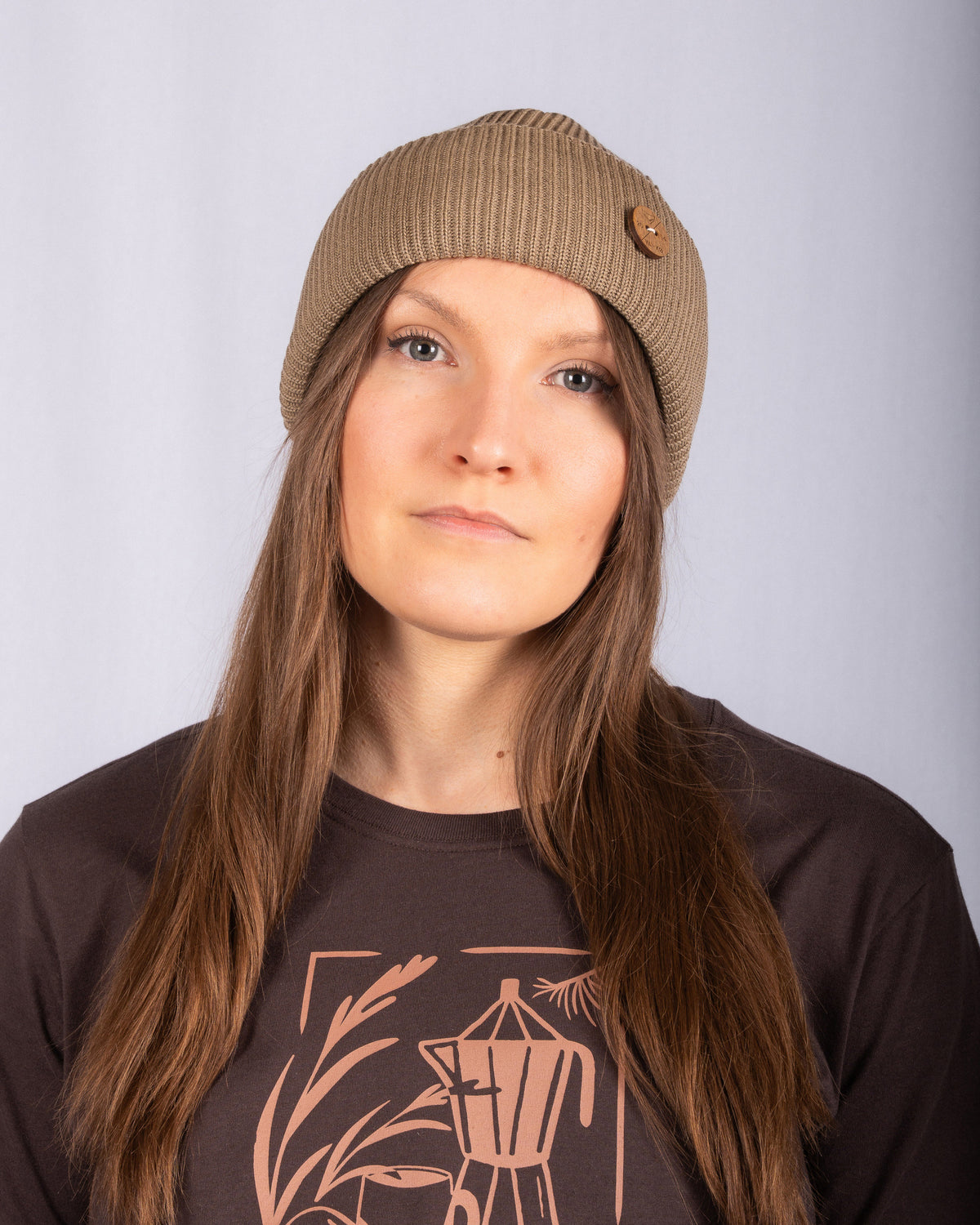 Woman with long brown hair wears a tan beanie and brown shirt with a coffee pot design, facing forward.