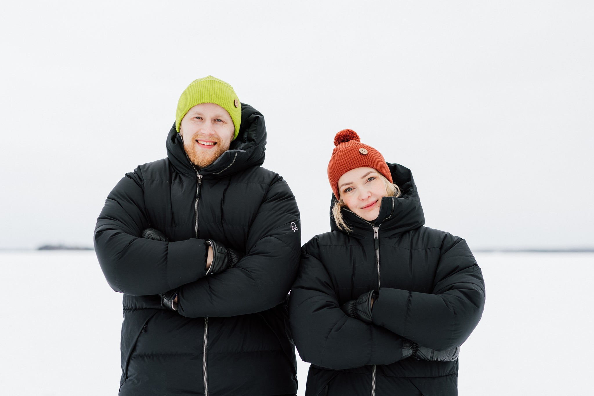 Two people in black winter jackets and colorful hats stand outdoors in a snowy landscape, smiling at the camera.