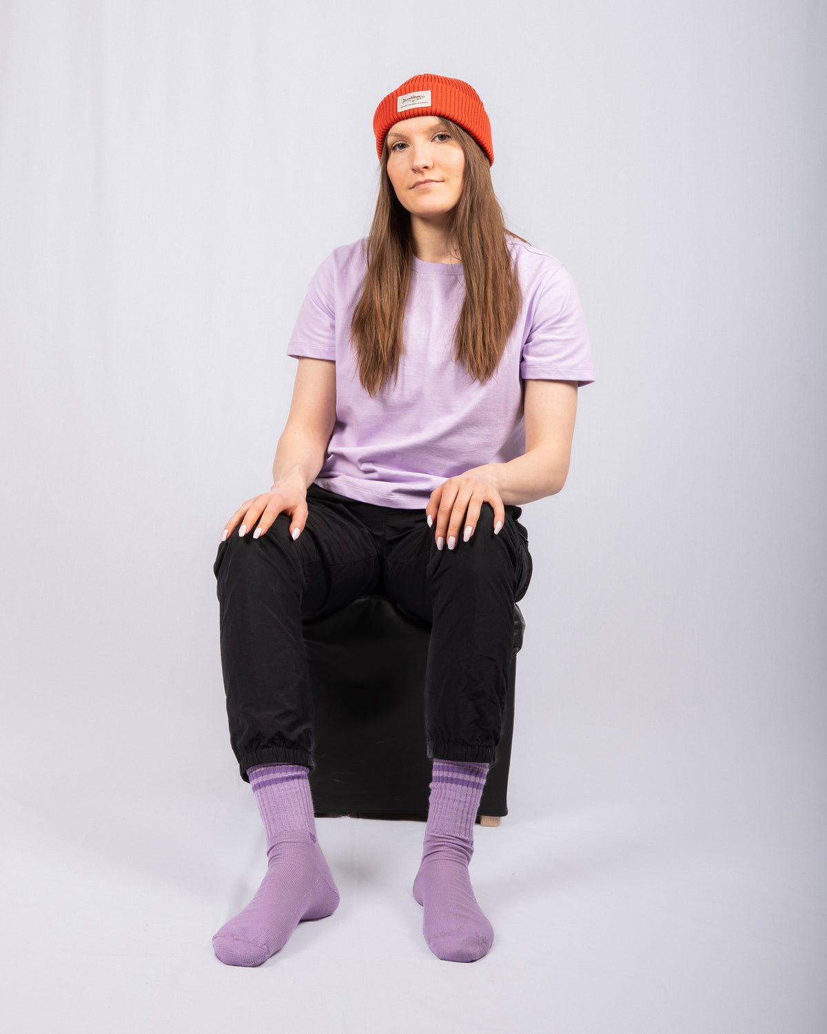 Person in lilac shirt, black pants, purple socks, and red beanie sits on a stool against a plain backdrop.