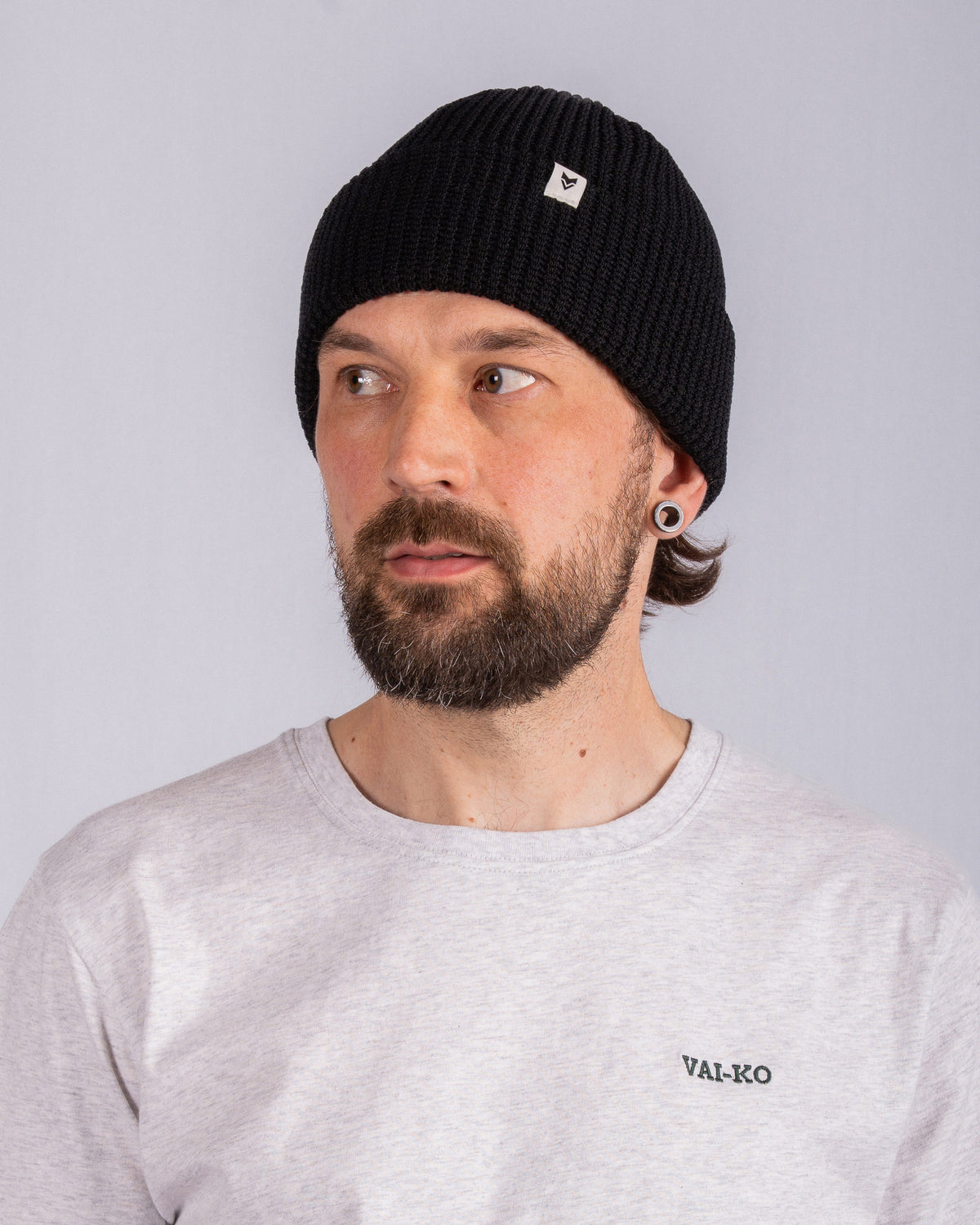 Man with beard wearing a black beanie and light gray VAI-KO shirt, looking to the side.