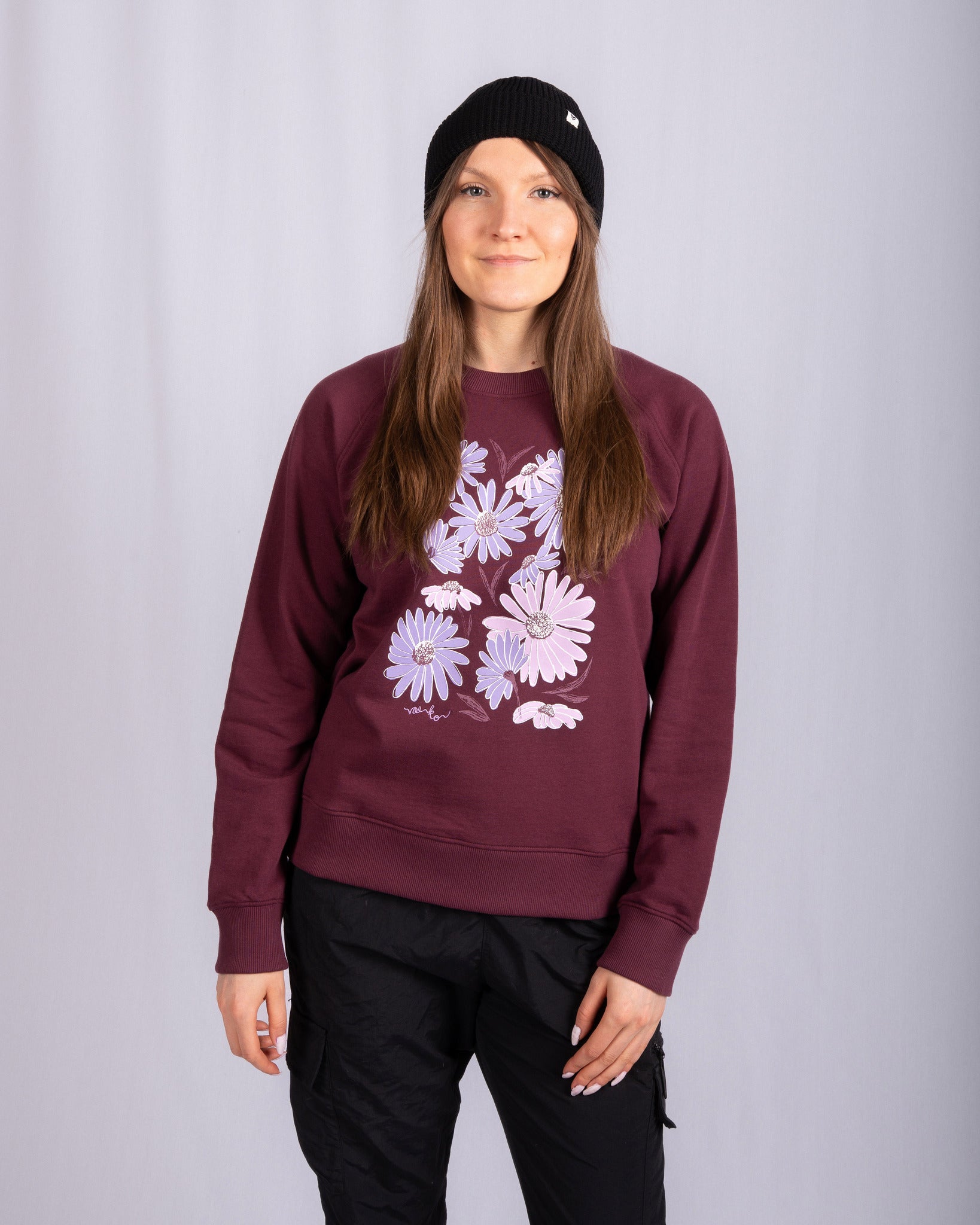 Woman in a maroon sweatshirt with purple flowers, black beanie, and black pants standing against a light backdrop.