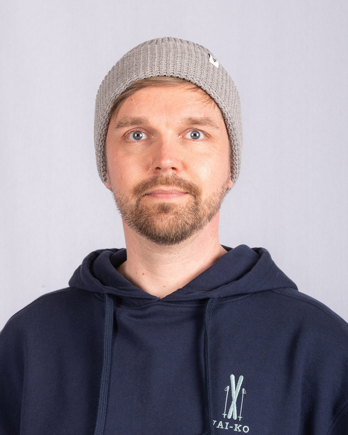 Man in gray knit beanie and navy hoodie stands against a plain light background, facing forward.
