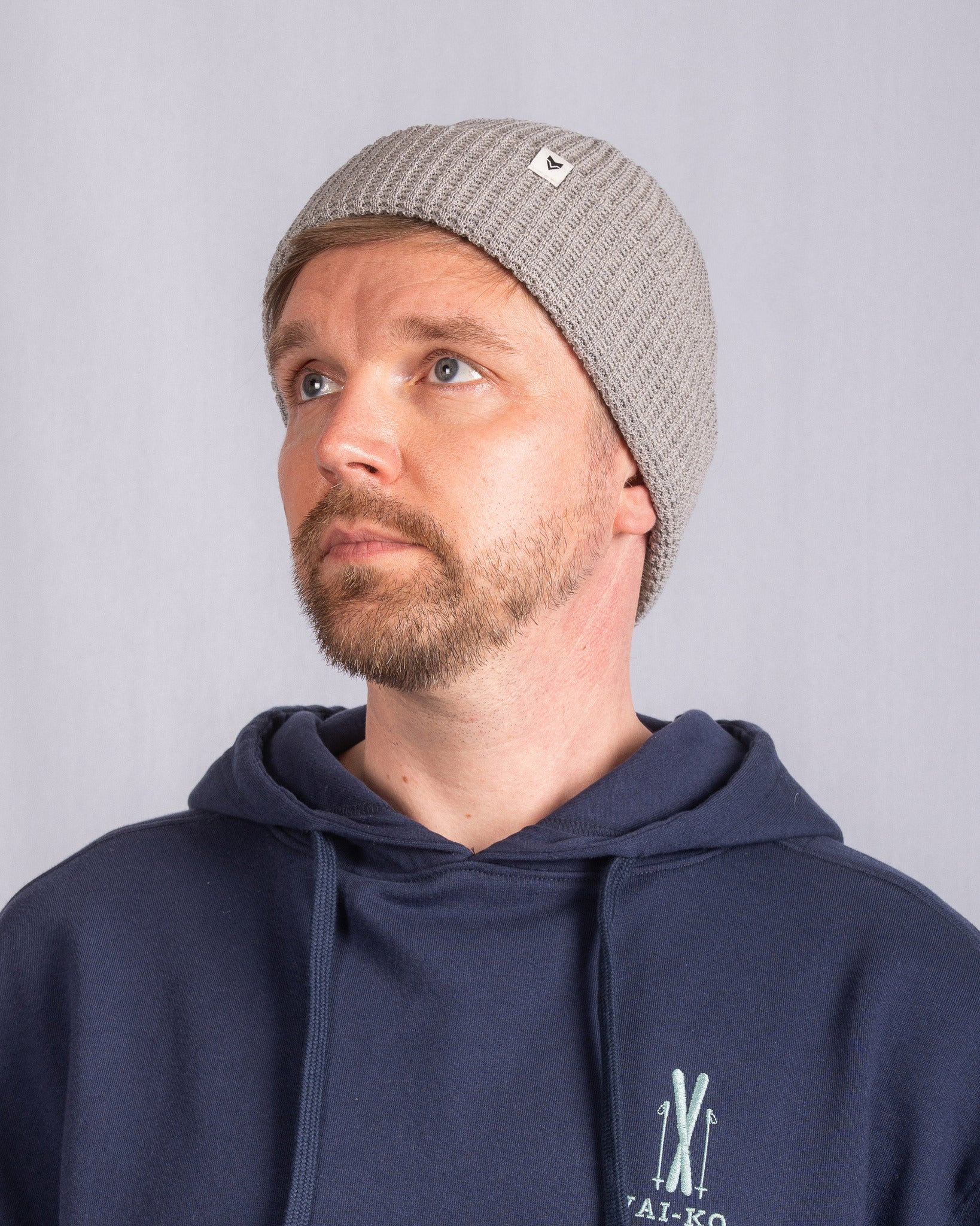Man in gray knit beanie and navy hoodie looks up, neutral background, white logo on beanie.