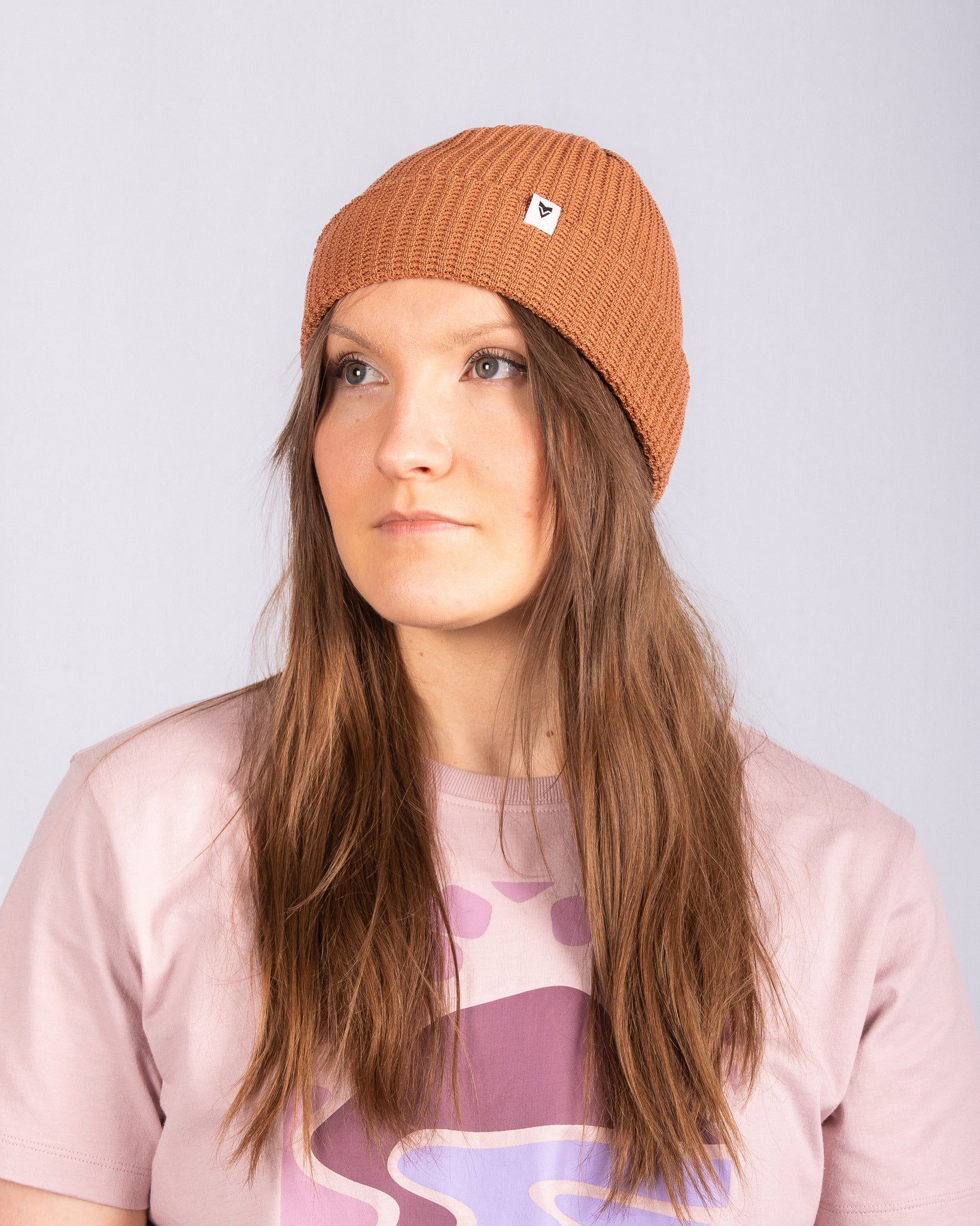Woman in a brown knit beanie and pink graphic t-shirt looks slightly to the left, against a plain background.