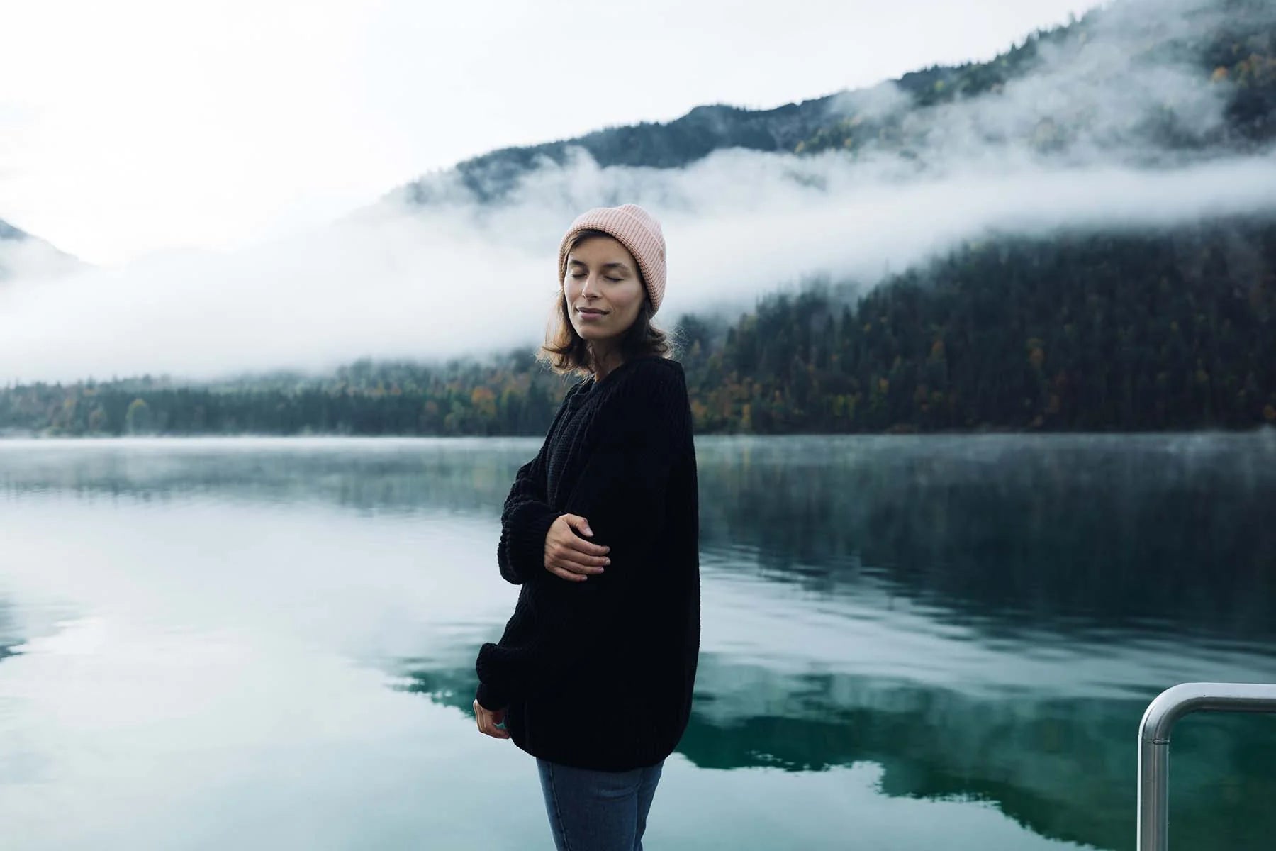 Woman in a pink beanie stands by a calm lake with misty mountains in the background.