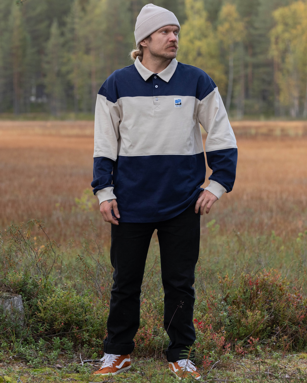 Man in a beanie and color-blocked shirt stands outdoors on grassy field with trees in the background.