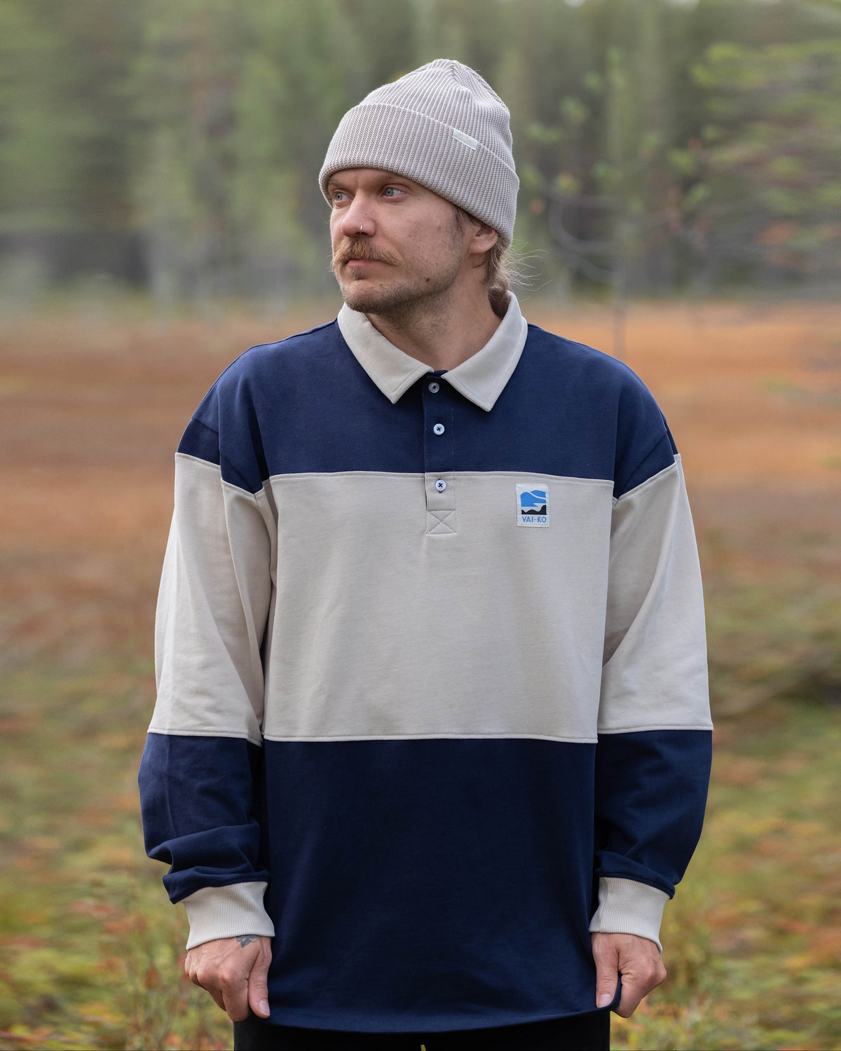 Man in a beige beanie and navy-and-beige pullover stands outdoors with a blurred forest background.